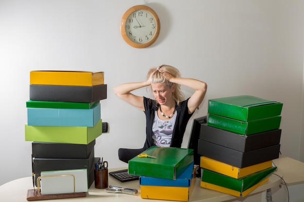 A teacher sitting at her desk, looking stressed and overwhelmed, with piles of papers and books surrounding her. The lighting is dim, and the overall feeling is one of exhaustion and pressure.