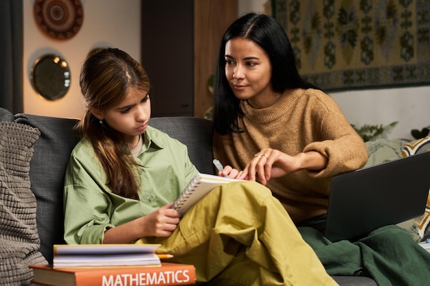 A young girl working on a math problem with a tutor in a classroom setting. The tutor is pointing at the problem and smiling encouragingly. They are surrounded by math textbooks and learning materials.