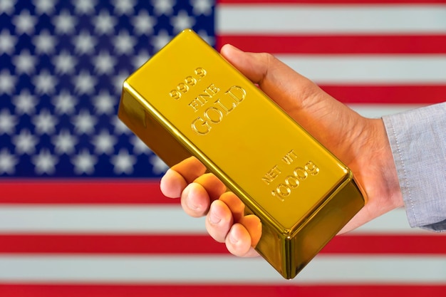 A close-up of a hand holding a gleaming gold bar, with the American flag subtly visible in the background, symbolizing the security and potential wealth associated with investing in precious metals.