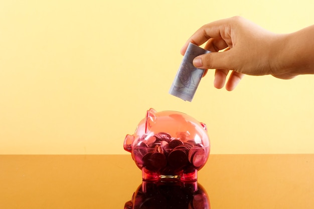 A close-up of a hand holding several coins of different denominations, with a piggy bank slightly out of focus in the background. The coins are stacked in small piles, suggesting the accumulation of savings over time. The overall image evokes the idea of thriftiness and the importance of minimizing investment costs.