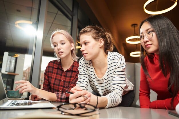 A female engineer mentoring a group of female high school students in a coding workshop session. Laptops and coding equipment are on the table, and the mentor is explaining a concept with enthusiasm.