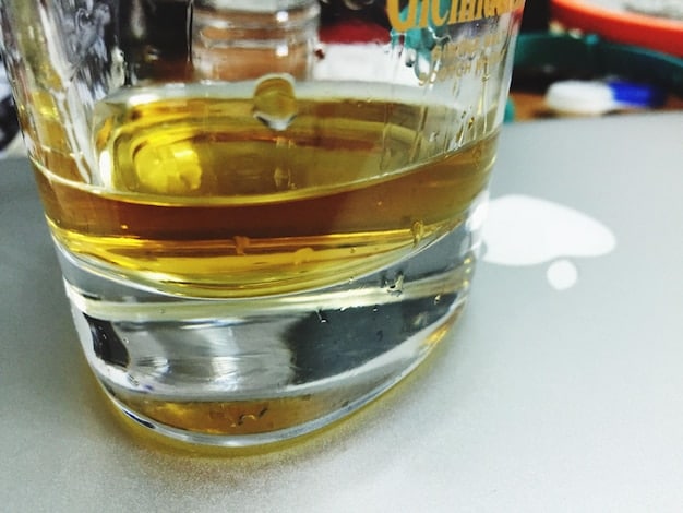 A close-up shot of biodiesel being poured from a beaker into a fuel tank. The biodiesel has a clear, golden hue. The fuel tank is labeled with warnings and specifications for biodiesel blends. The background shows a workshop with various tools and equipment used for fuel production and testing.